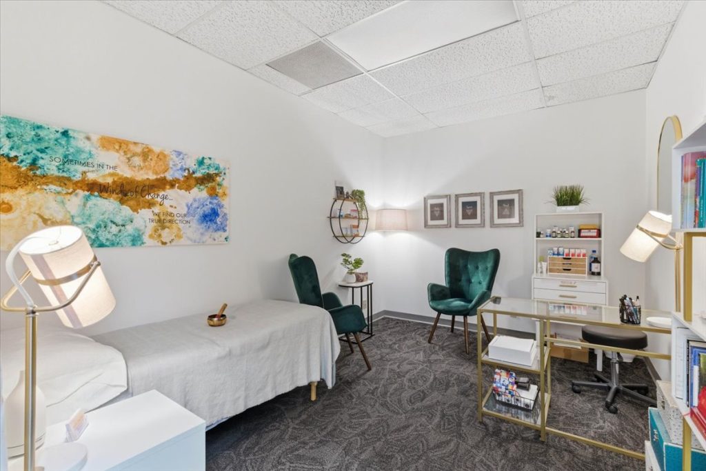 Homeopathy treatment room with a treatment table, green armchairs and product shelving